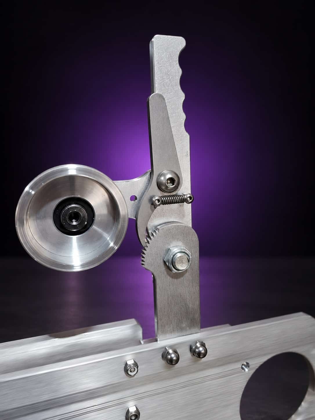 Classic grinder tension lever with spring and ratchet teeth beside the idler wheel, close-up, on black with a purple glow