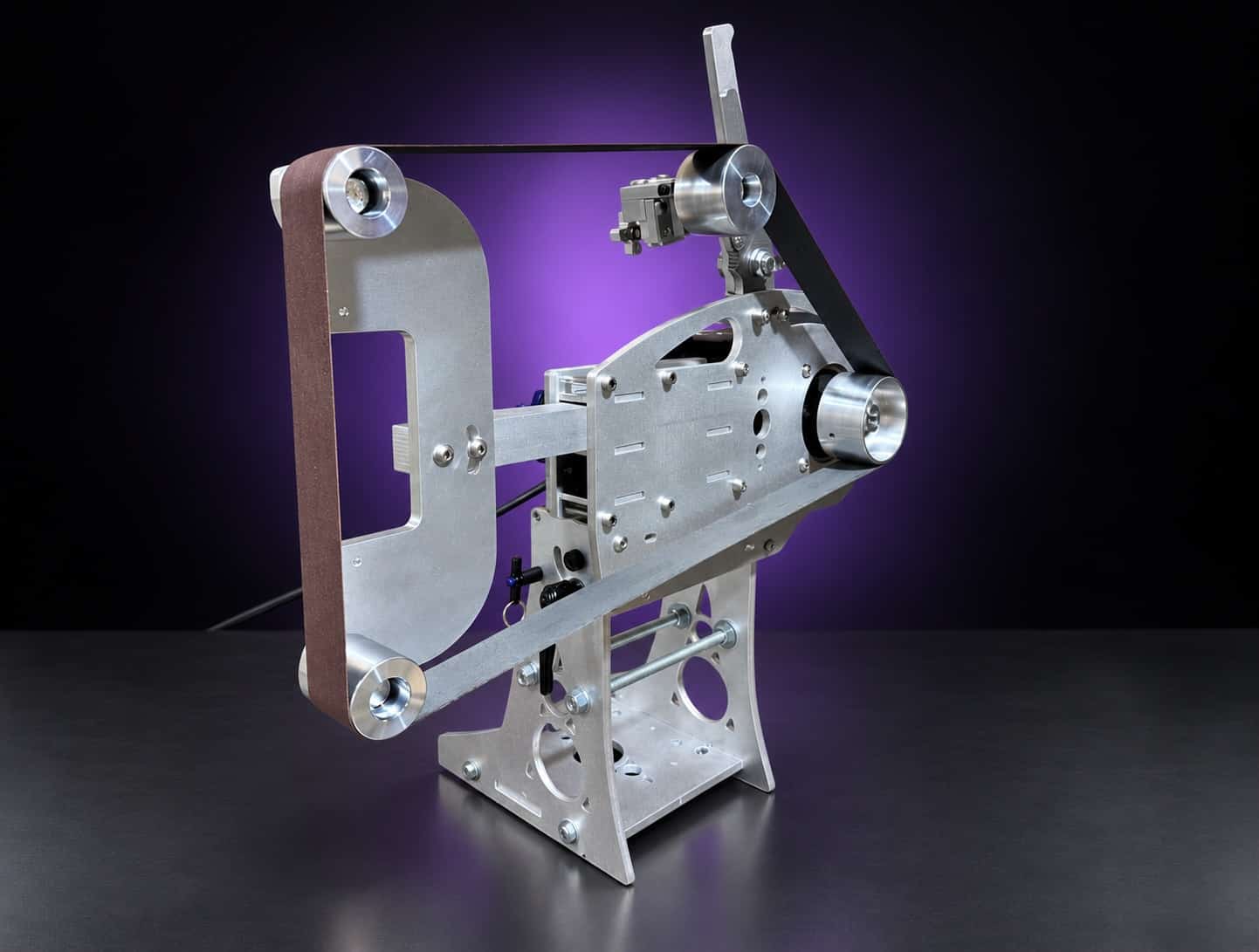 Nova aluminum 2x72 grinder with flat-platen tool arm and abrasive belt, three-quarter view, on black with a purple glow