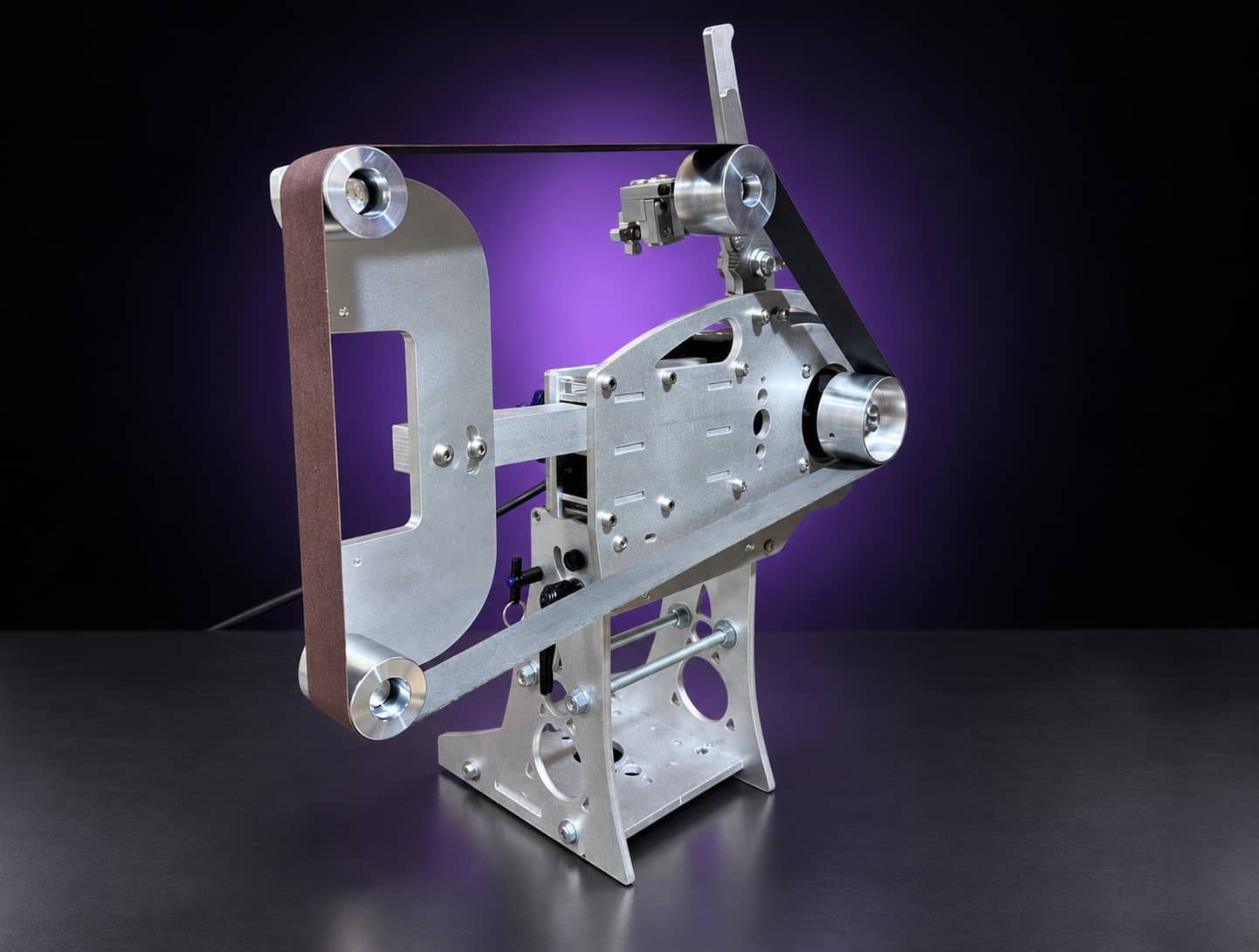 Nova aluminum 2x72 grinder with flat-platen tool arm and abrasive belt, three-quarter view, on black with a purple glow