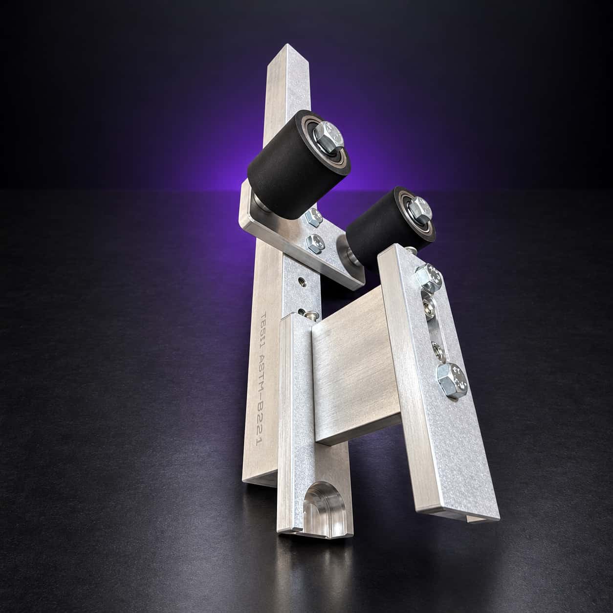 Small wheel attachment standing on end: aluminum arm, holder plate and two black rubber deflector wheels, on black with a purple glow